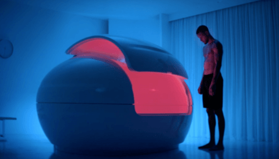 Guy standing next to sensory deprivation tank as it's opening the pod door and shining an orange-red light from inside.
